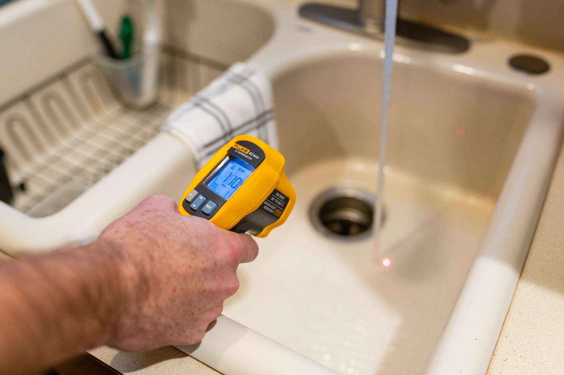 Hand holding a yellow infrared thermometer over a kitchen sink with running water