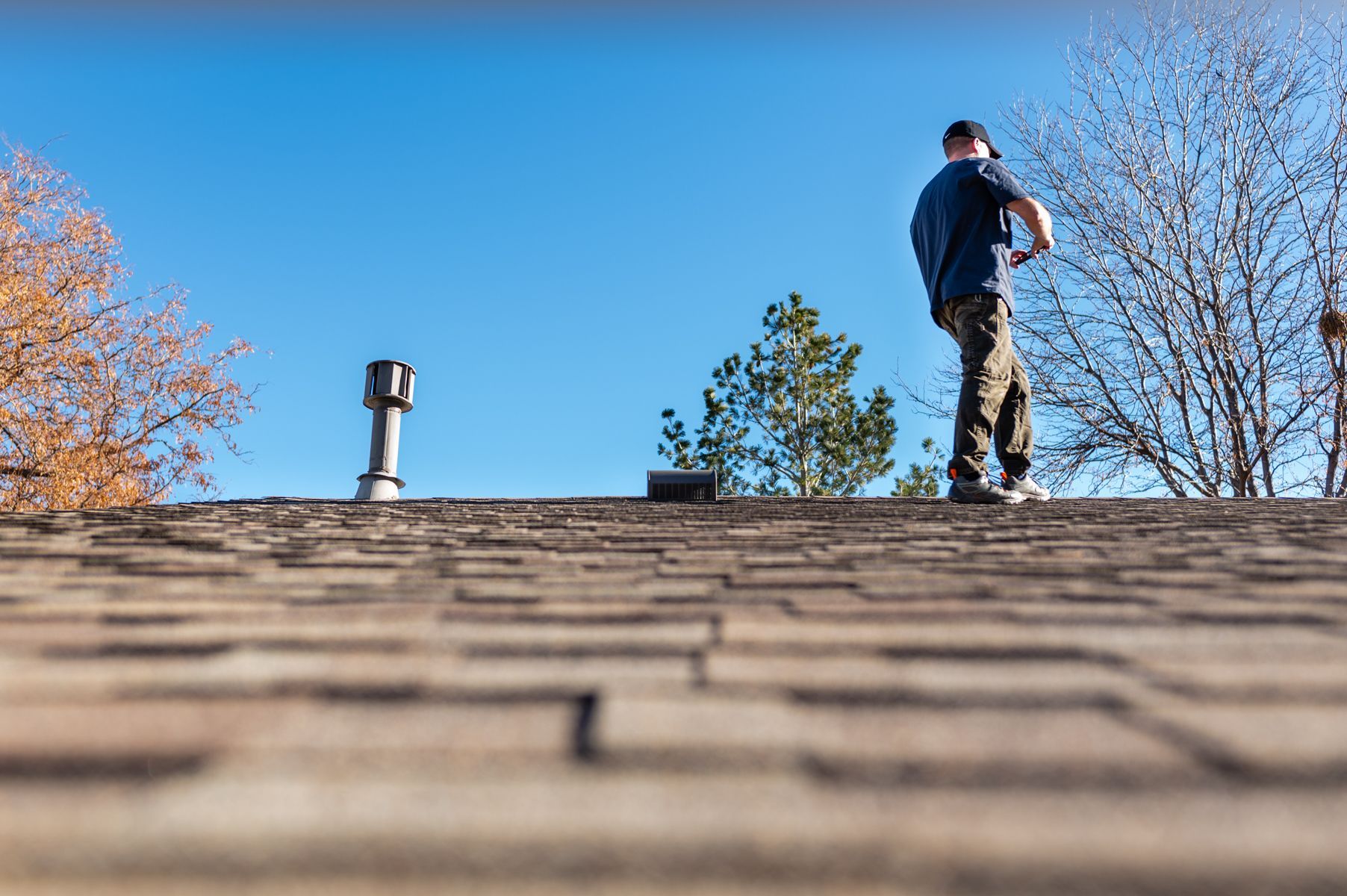 Person standing on a shingled roof under a clear blue sky, with trees and a chimney in the background