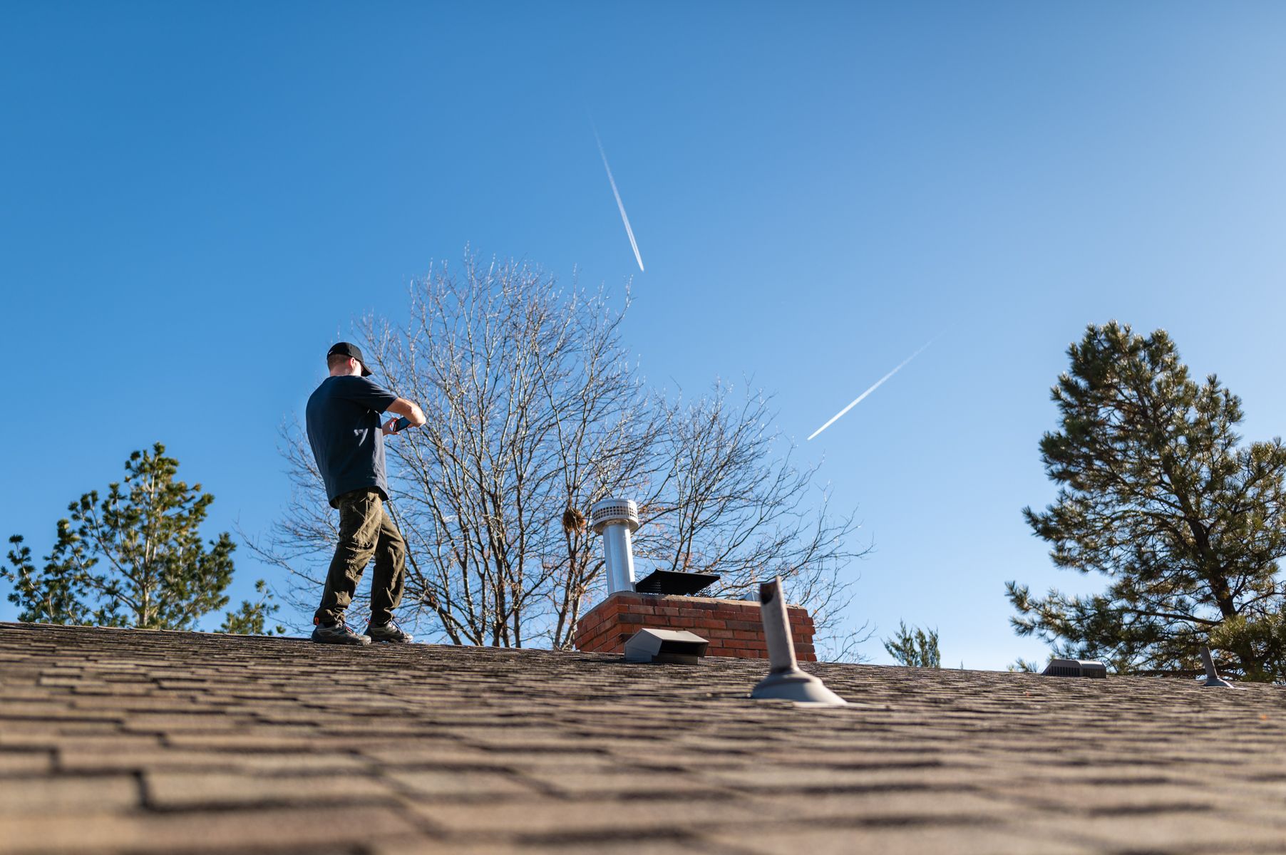 Person standing on a shingled roof aiming at the sky with a baseball bat, trees and blue sky behind.