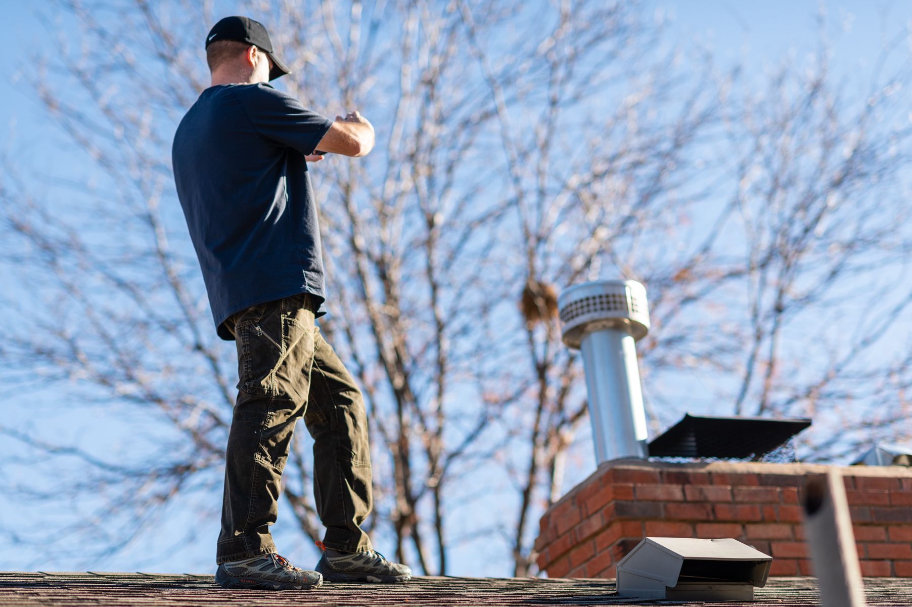 Person standing on a rooftop beside a chimney, looking out over bare trees on a clear day