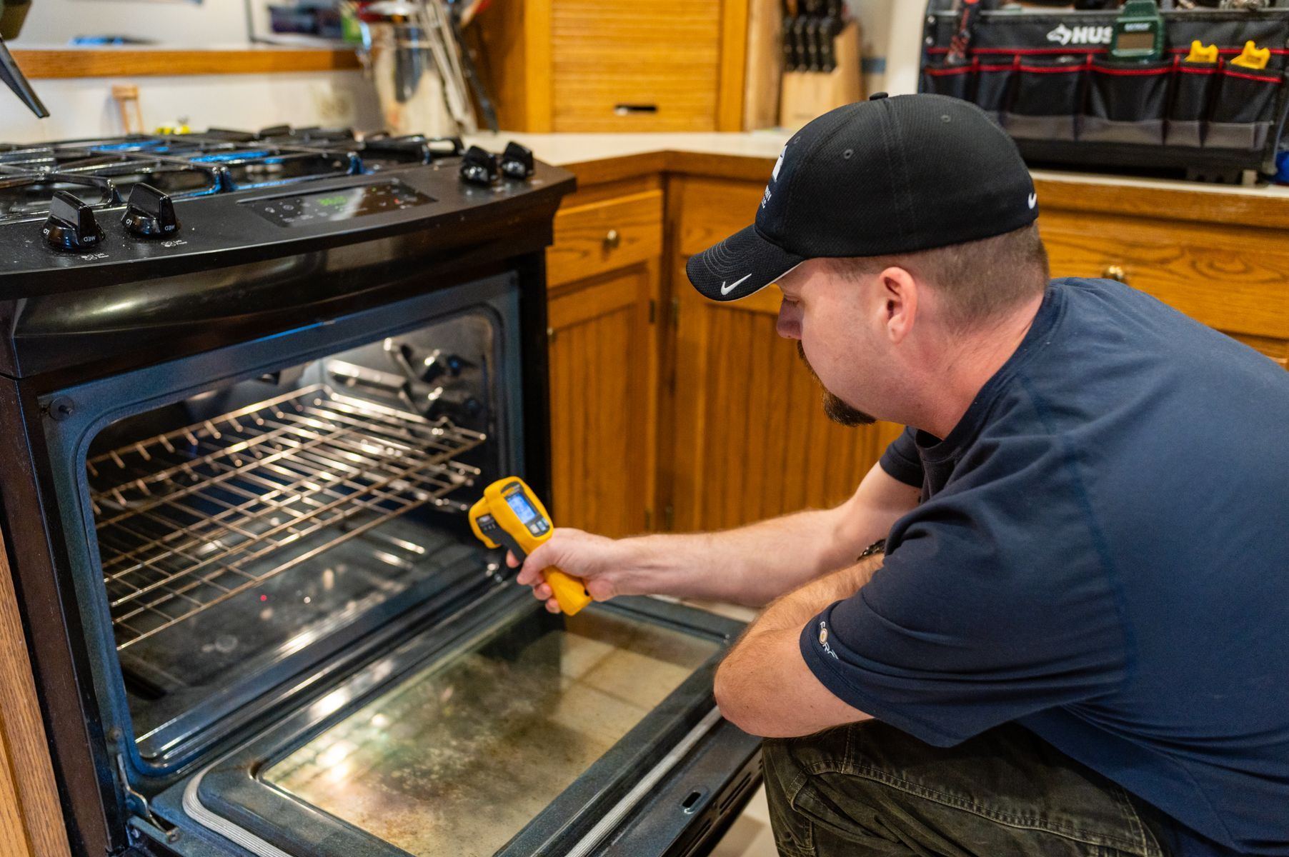 Man cleaning a kitchen oven door with a yellow tool while kneeling beside the open oven