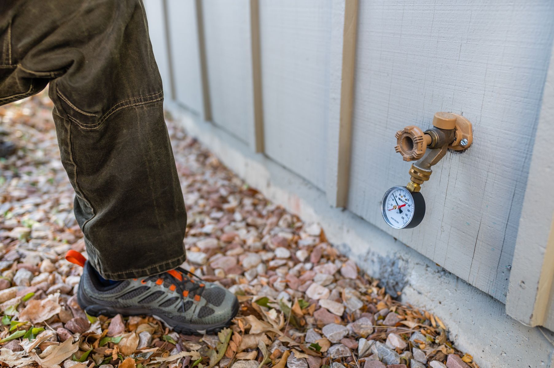 Person standing by an outdoor water spigot with a pressure gauge on a shed wall
