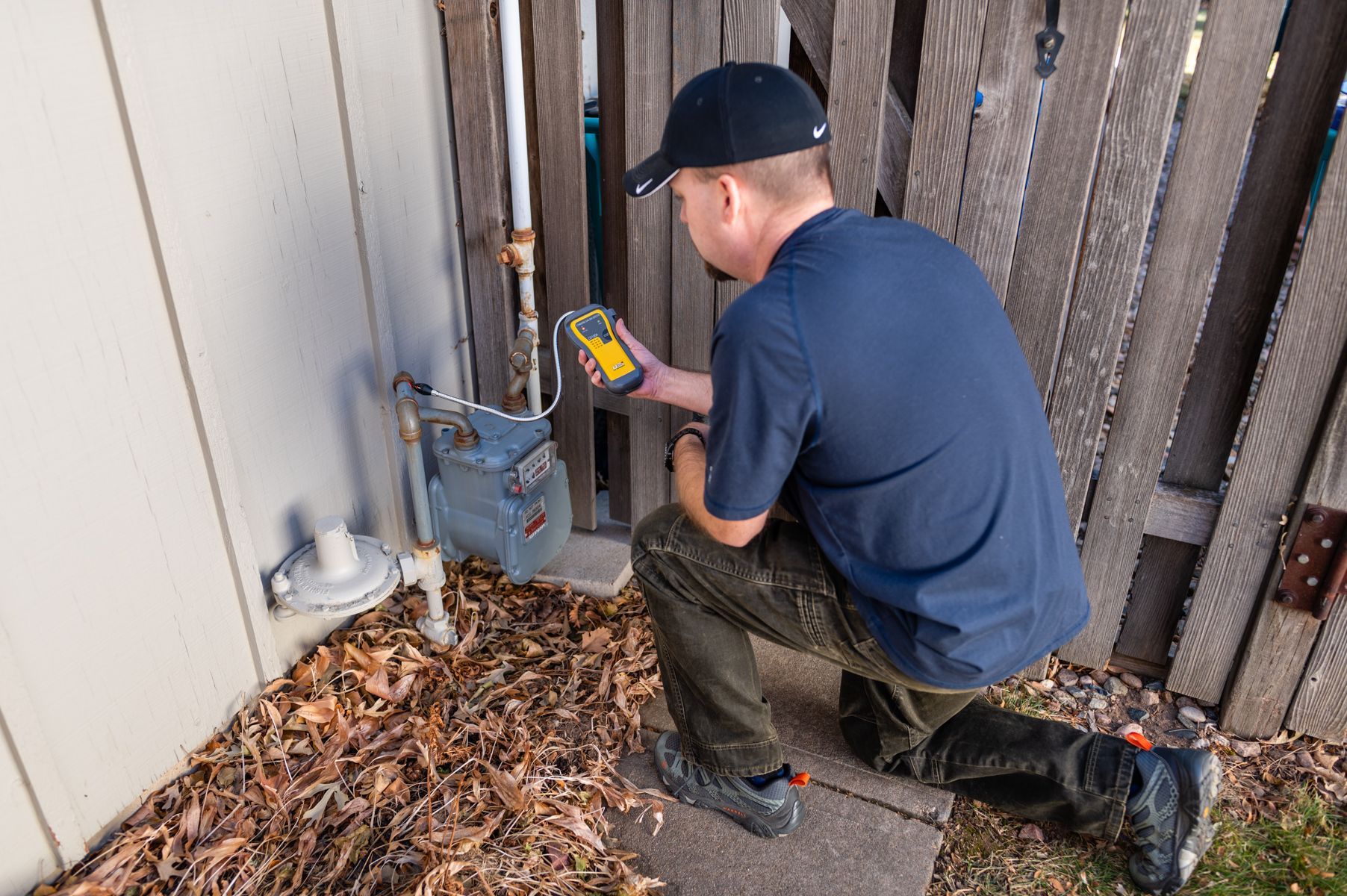 Worker kneeling by an outdoor utility box, using a handheld tester beside a house wall and fence.