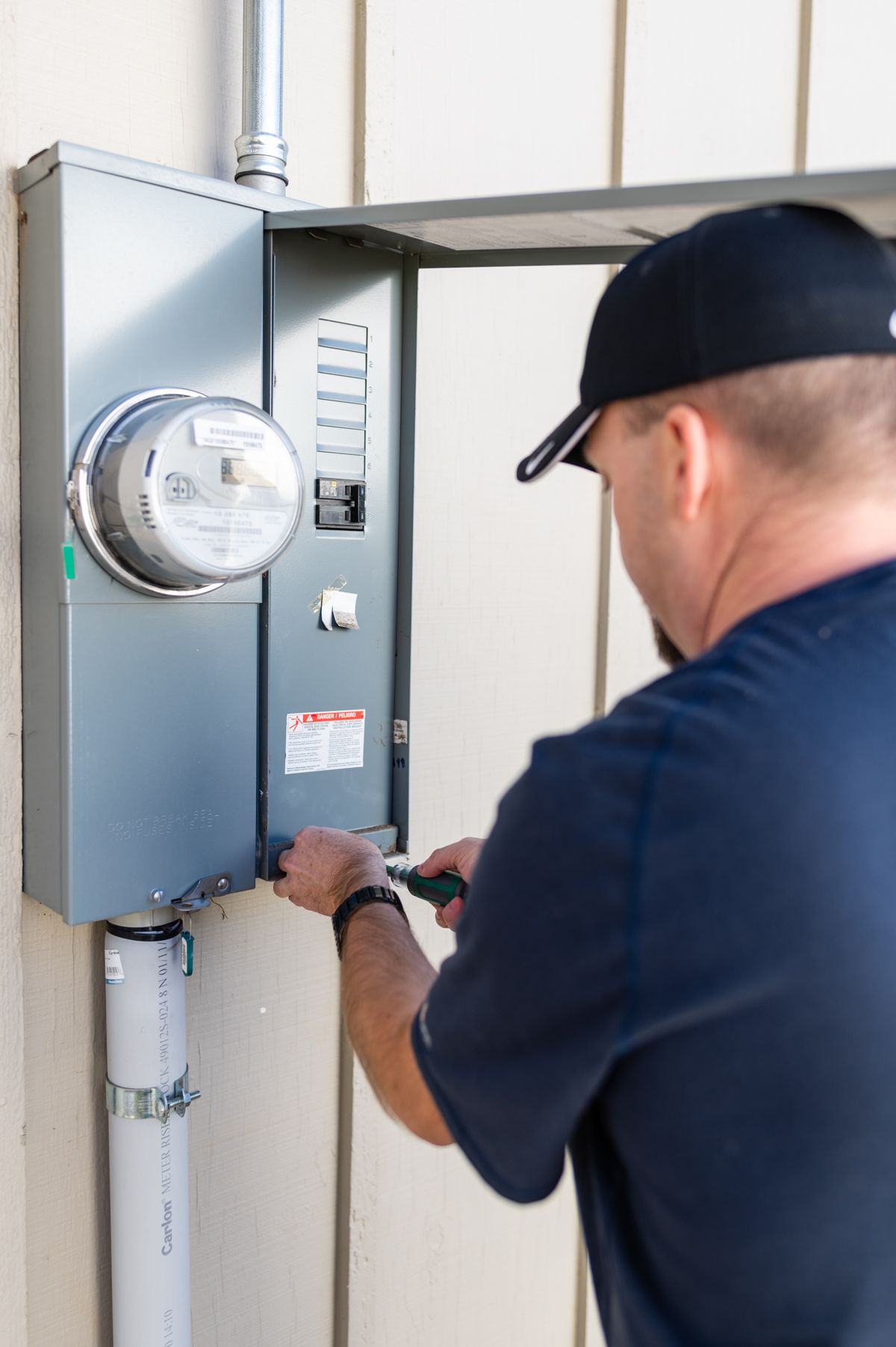 Electrician working on an open outdoor electrical meter panel beside a gray wall