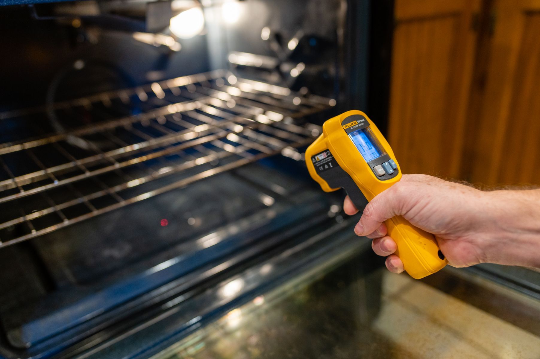 Hand holding a yellow infrared thermometer aimed at a hot oven interior in a kitchen