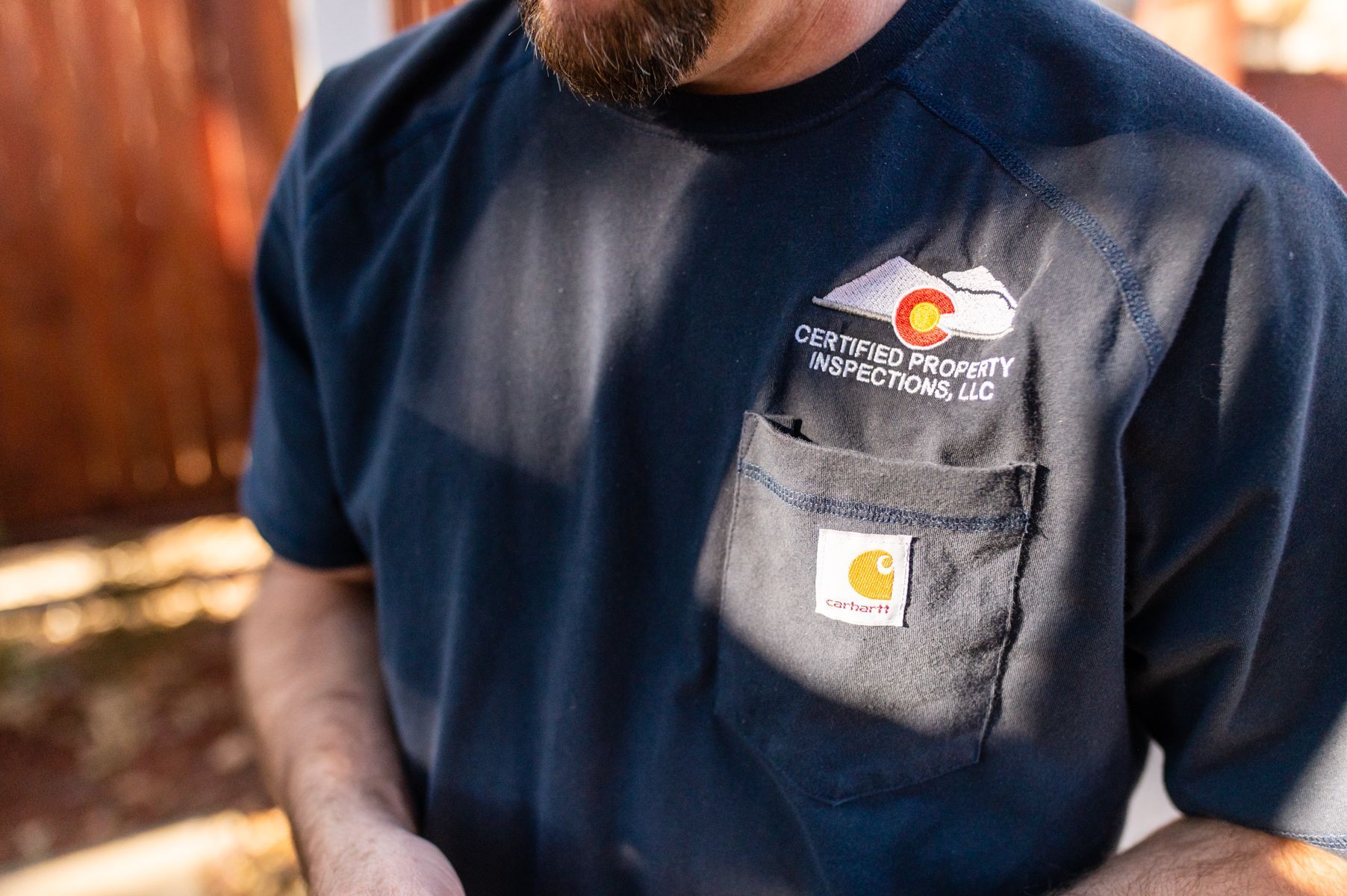 Man in navy T-shirt with logo and chest pocket, standing outdoors in warm sunlight