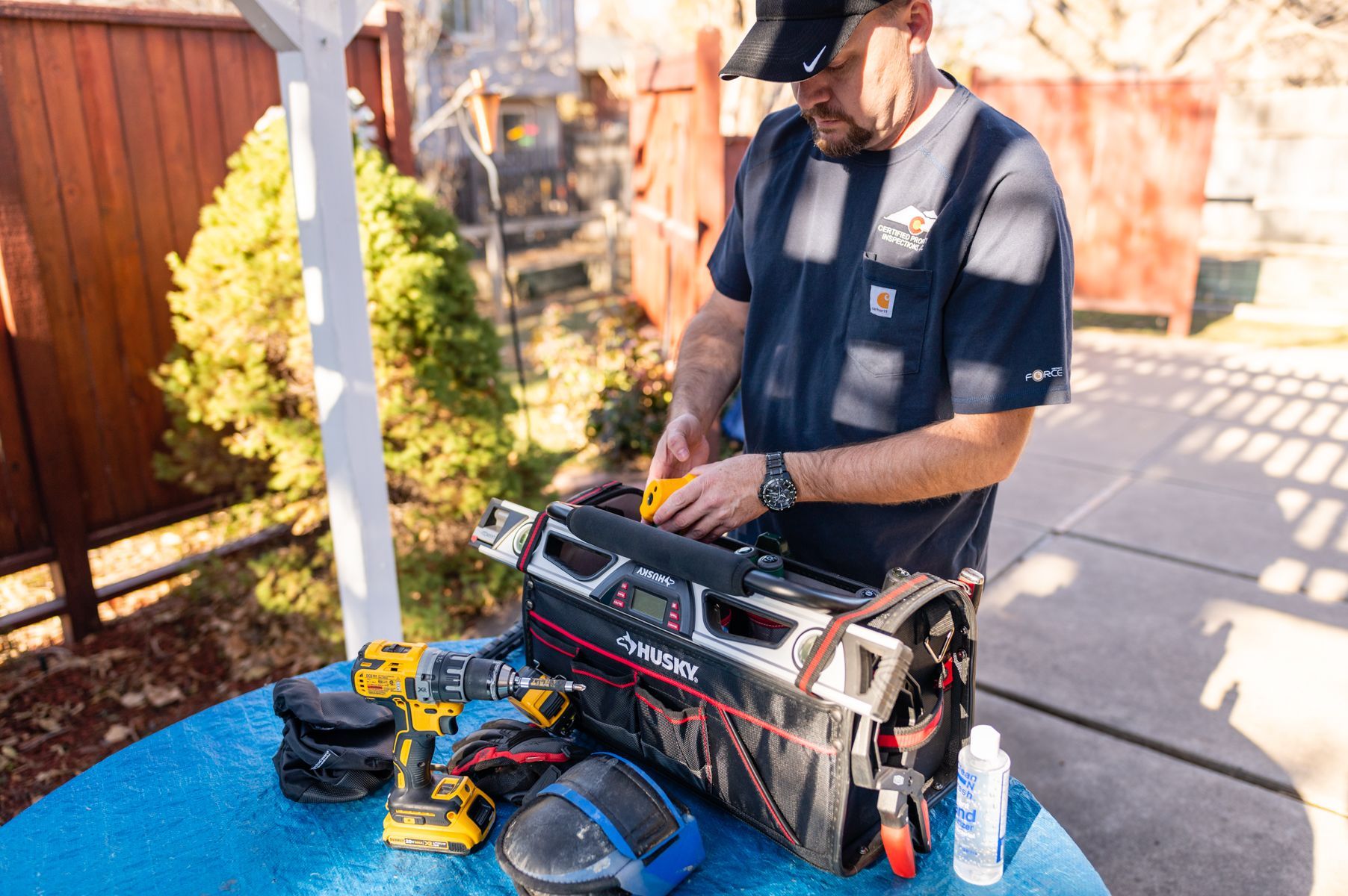 Worker inspecting tools on a blue table in a backyard, with a toolbox and power drills nearby.