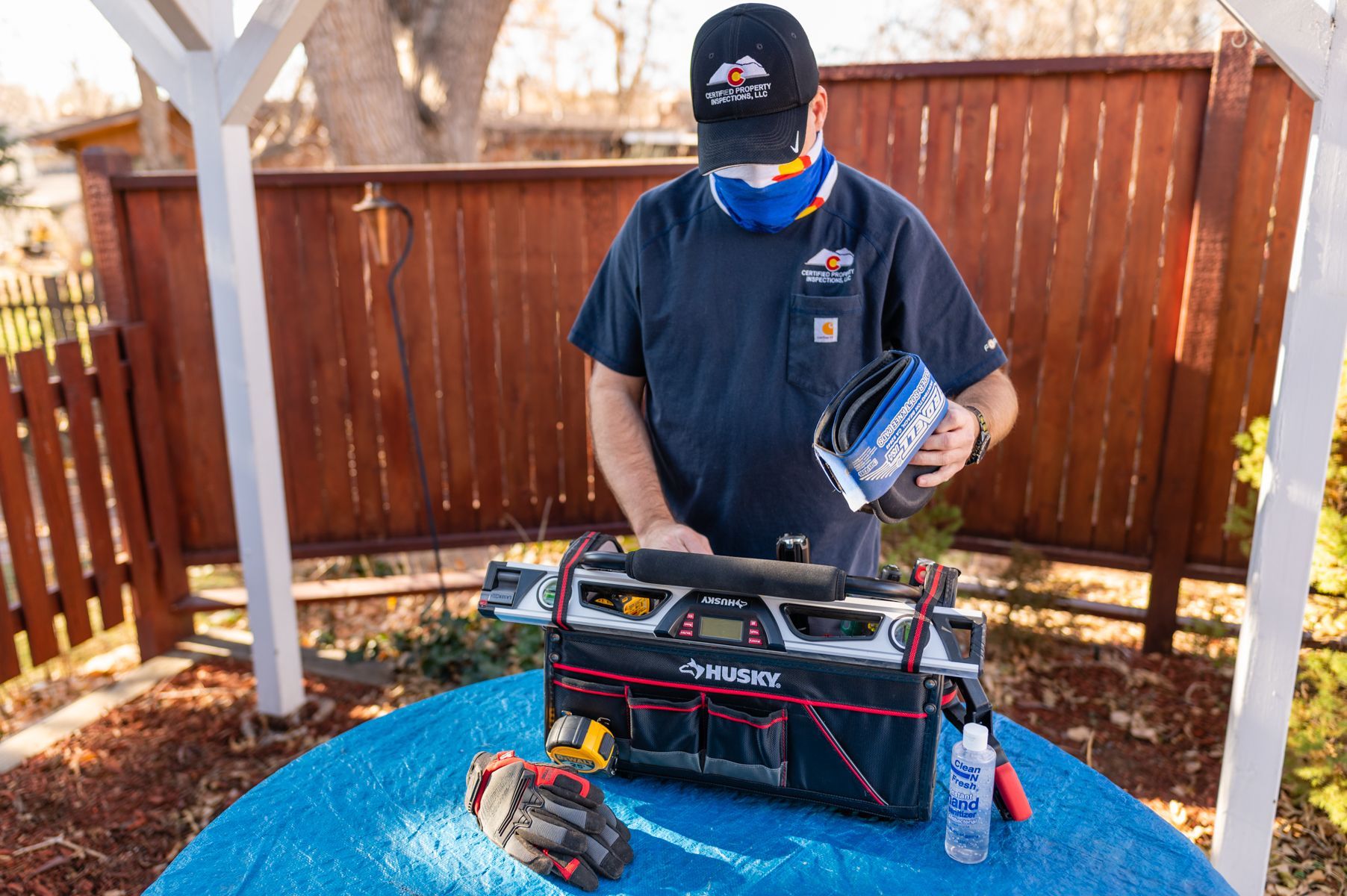 Person in a backyard holding a tool over a table with tools and supplies, wearing a mask and gloves.