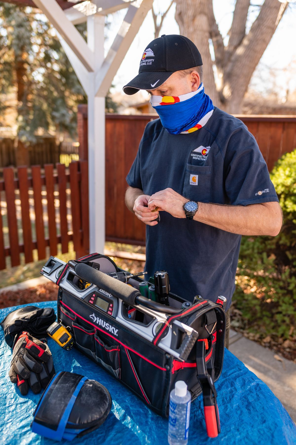 Person in a cap and blue mask adjusts a tool bag on a blue table outdoors.