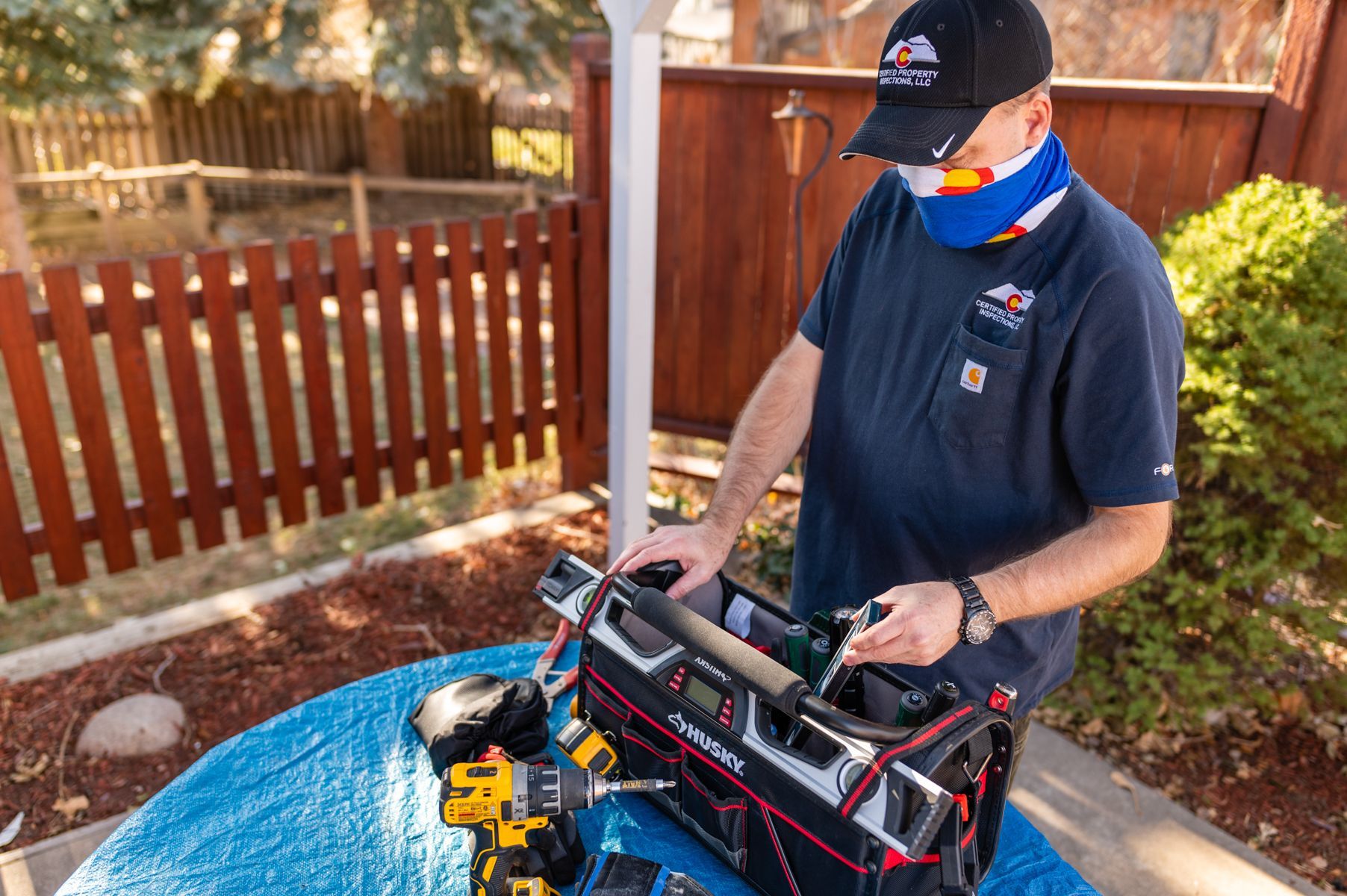 Man in navy shirt and cap working on tools at a patio table beside a wooden fence