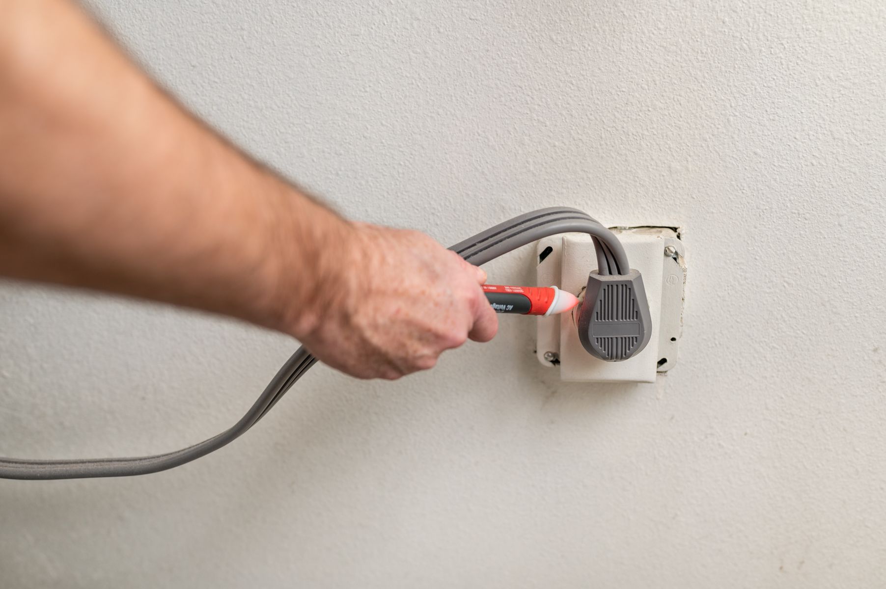 Hand plugging a gray power cord into a wall outlet with a red tester tool