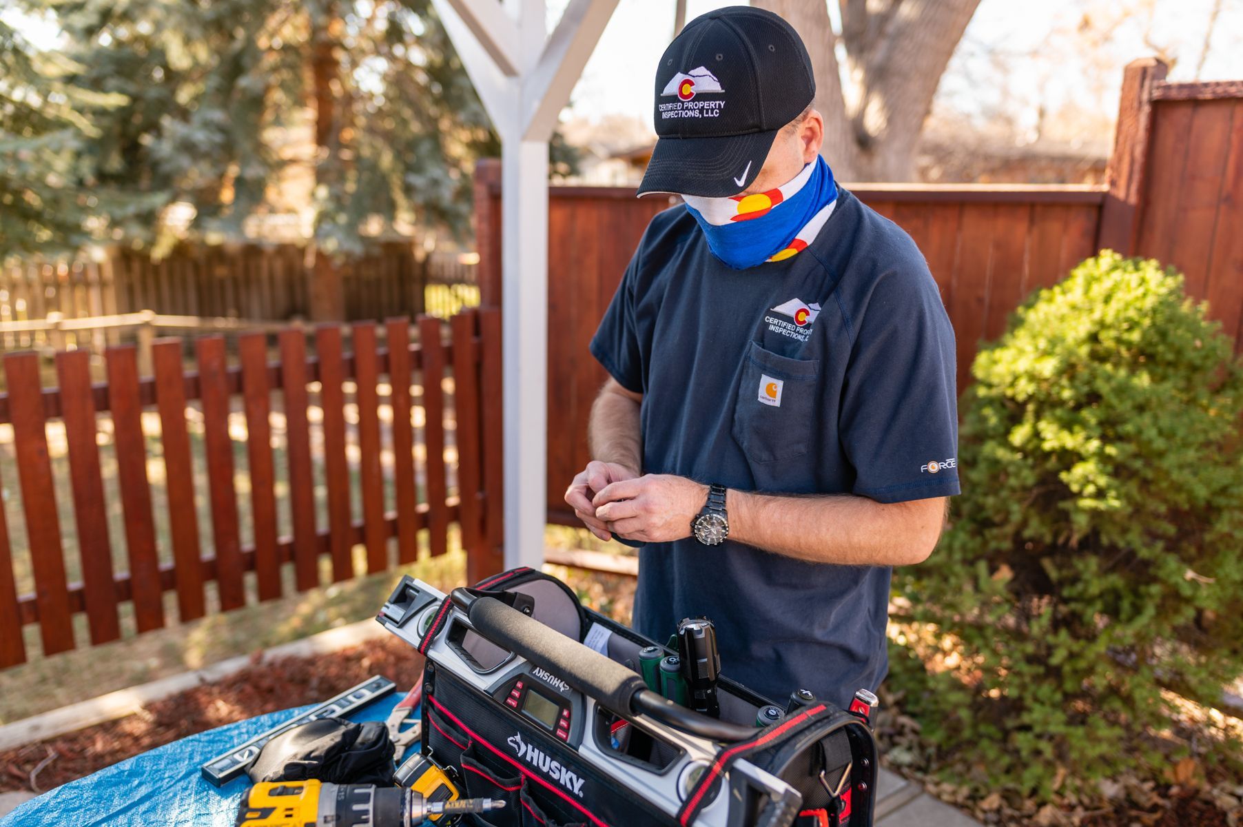 Person outdoors working at a bench, wearing a cap and mask, with tools on the table in a backyard.