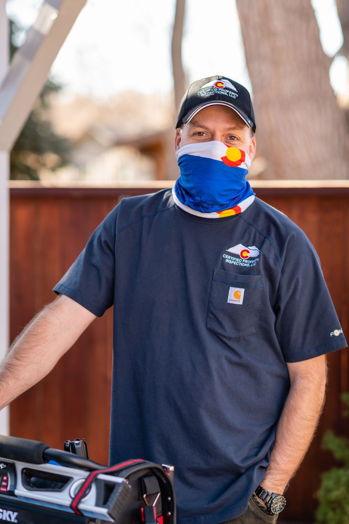 Person wearing a navy cap, navy shirt, and blue face covering, standing beside a red-and-black object outdoors.
