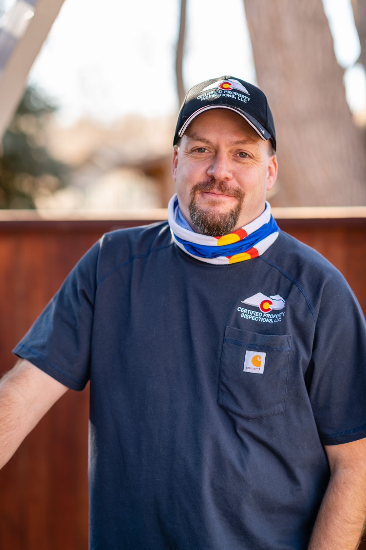 Man in a navy shirt and cap standing outdoors, wearing a colorful neck gaiter and smiling.