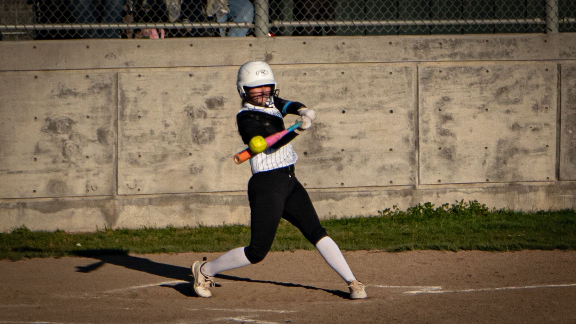 Softball player in black uniform swinging a bat on a dirt field in front of a concrete wall