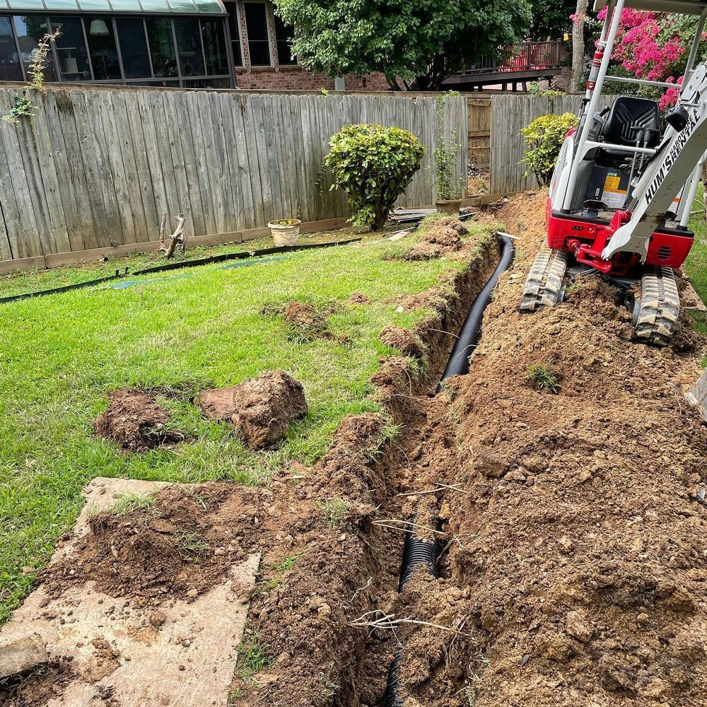 Mini excavator in a trench next to a black pipe in a backyard.