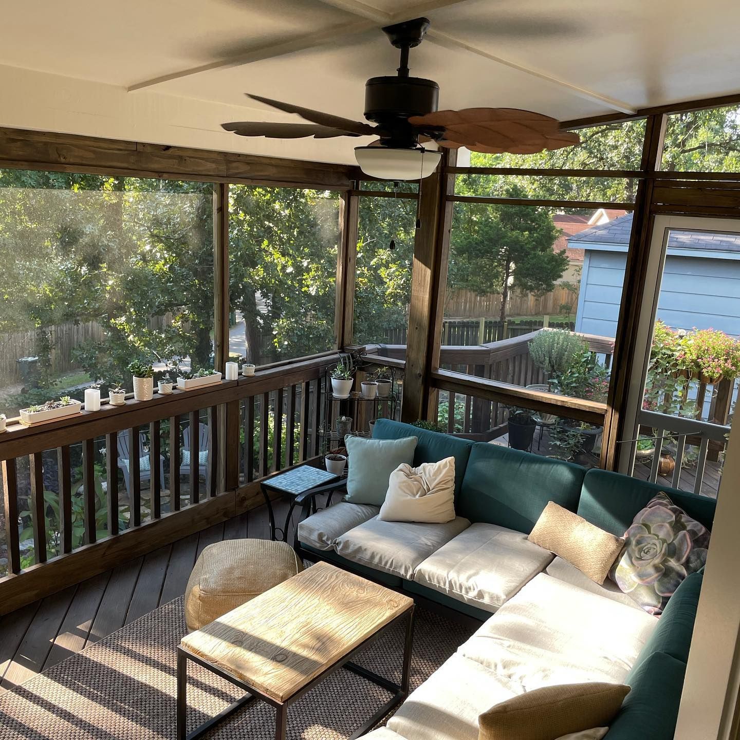 Screened-in porch with teal couch, wicker coffee table, wooden ceiling fan, and view of trees.