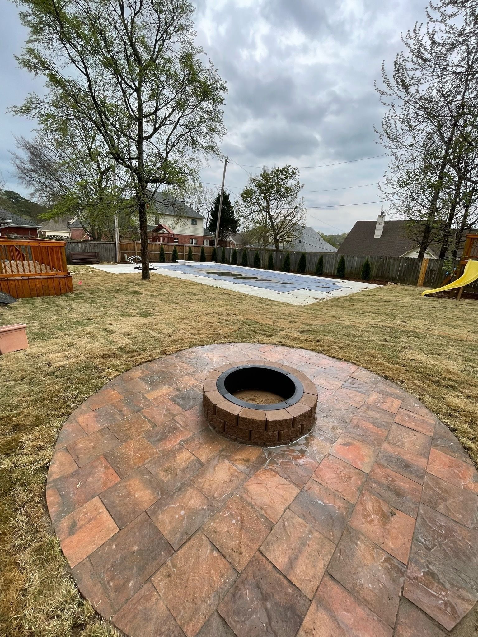 Brick fire pit in a backyard with a covered pool and overcast sky.