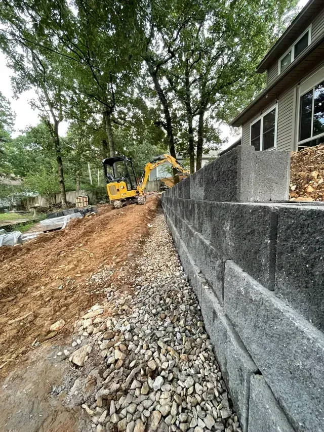 Construction equipment grading a dirt slope beside a house and stone retaining wall