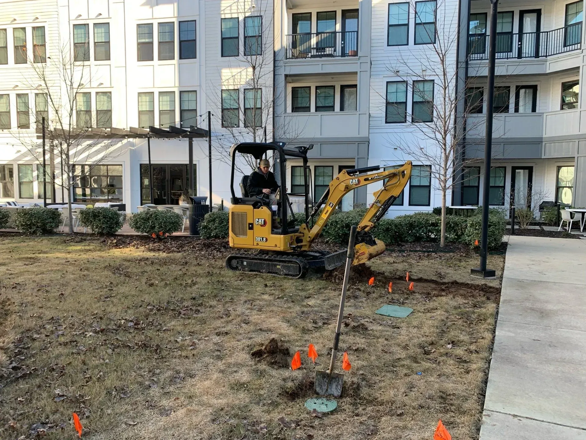 Mini excavator digging in a grassy area near an apartment building.