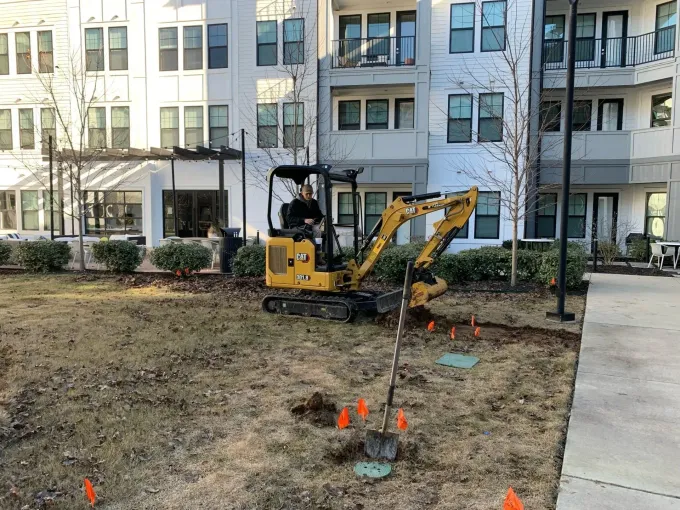 Yellow excavator working in a dirt courtyard beside an apartment building, with orange survey flags and a sidewalk nearby