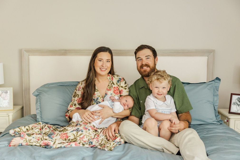 Family sitting on a bed with two young children, smiling in a softly lit bedroom.