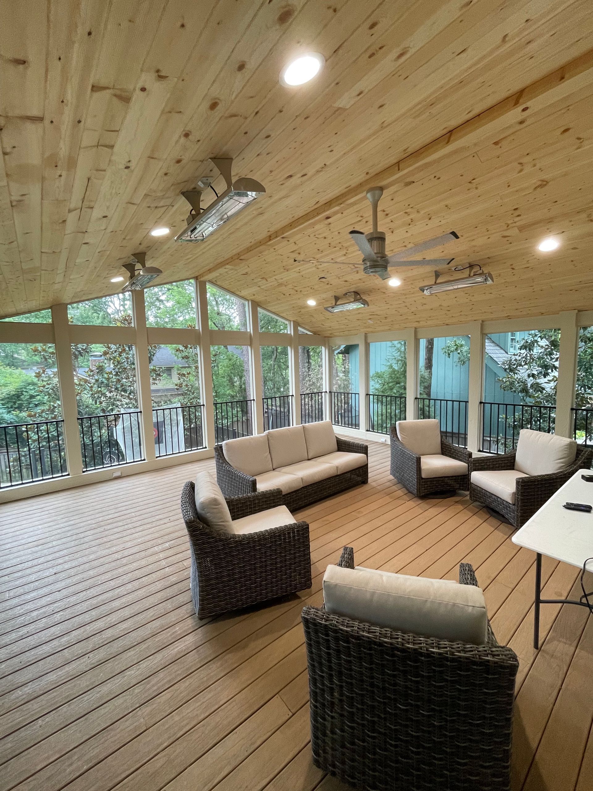 Covered outdoor living space with wood ceiling and floor, wicker furniture, and large windows overlooking trees.