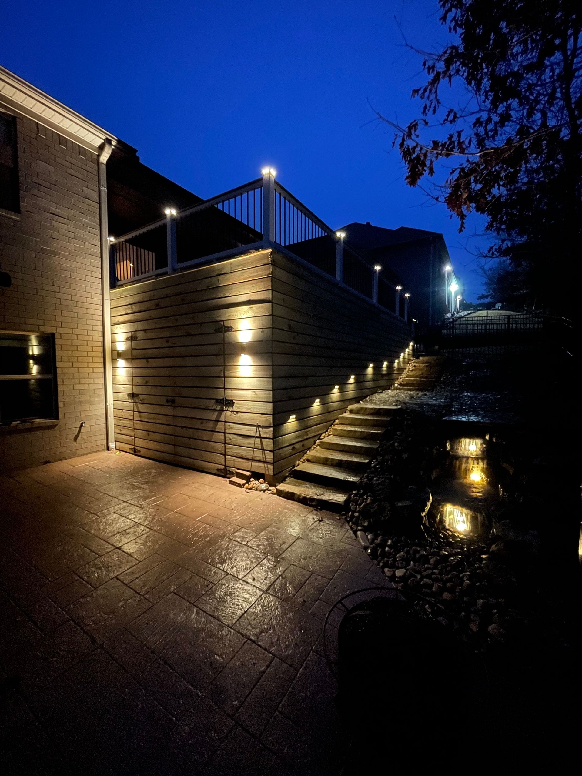 Exterior night scene of a house with steps and deck illuminated by outdoor lights. Dark blue sky.