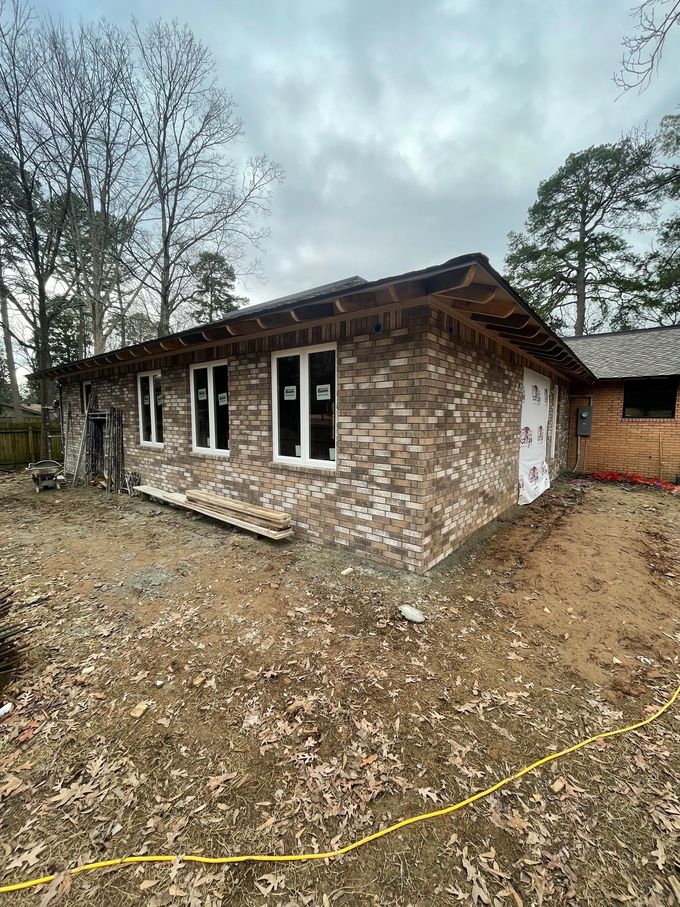 Single-story brick house under construction, with gravel yard, windows, and trees in the background.
