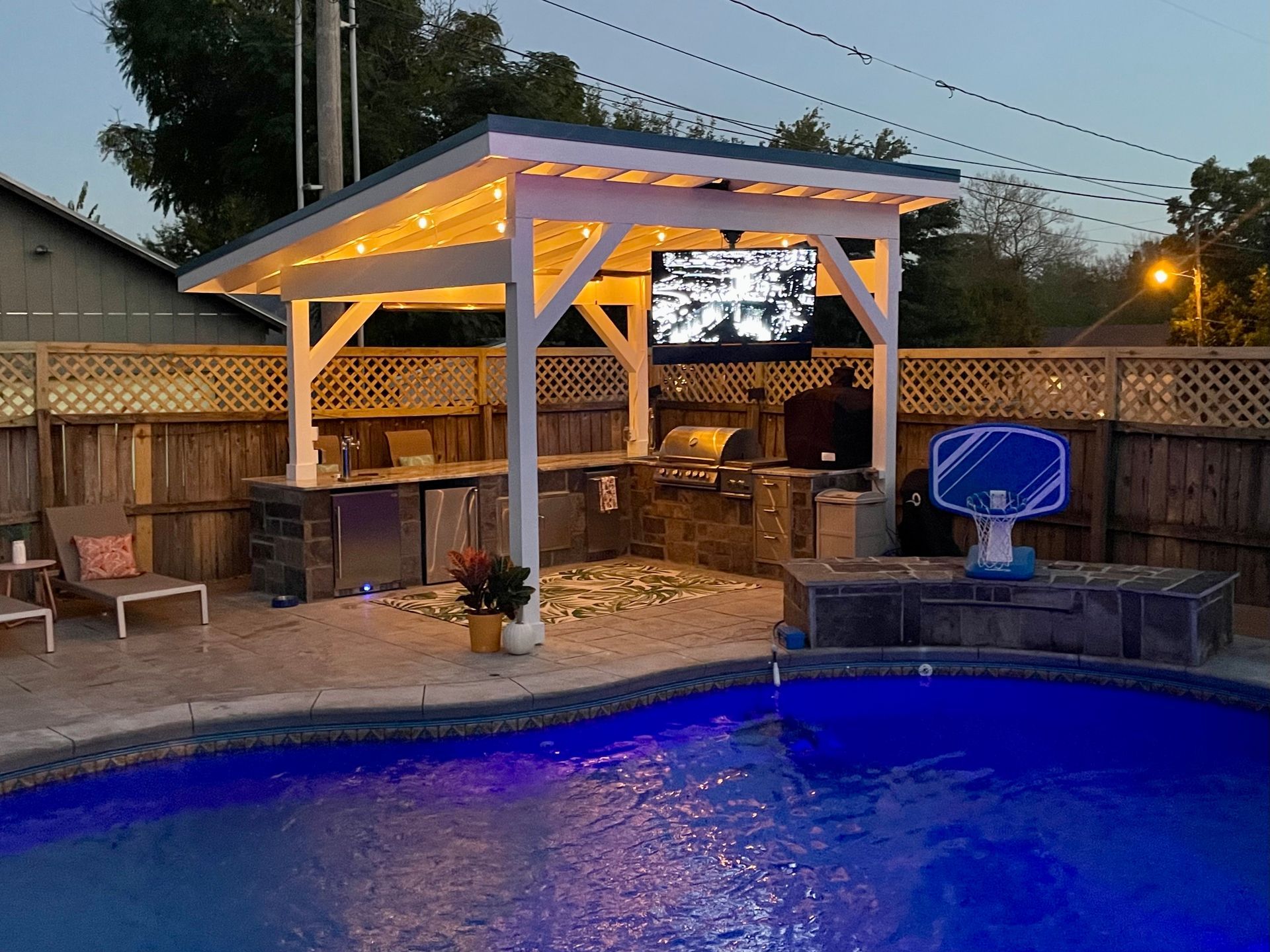 Backyard pool at dusk with lit gazebo, TV, fire pit, and basketball hoop