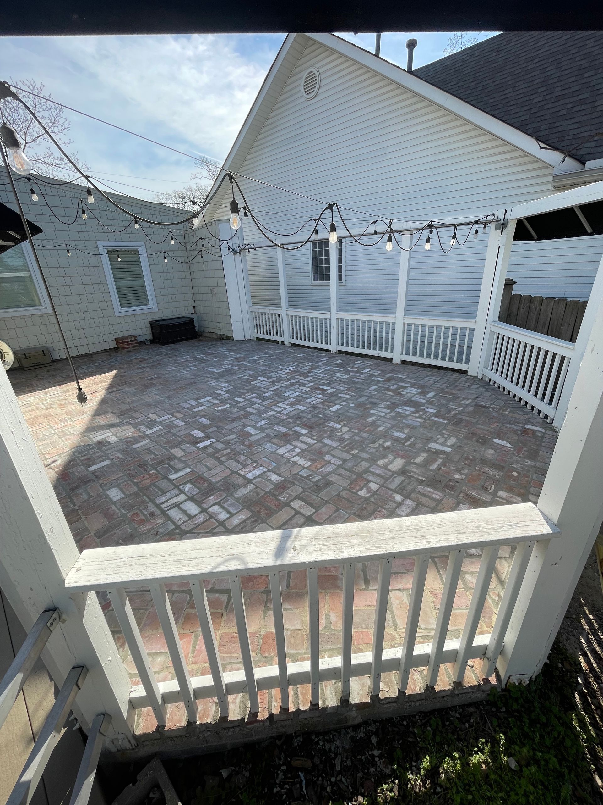 White fenced brick patio with string lights between white houses, viewed from a backyard deck.
