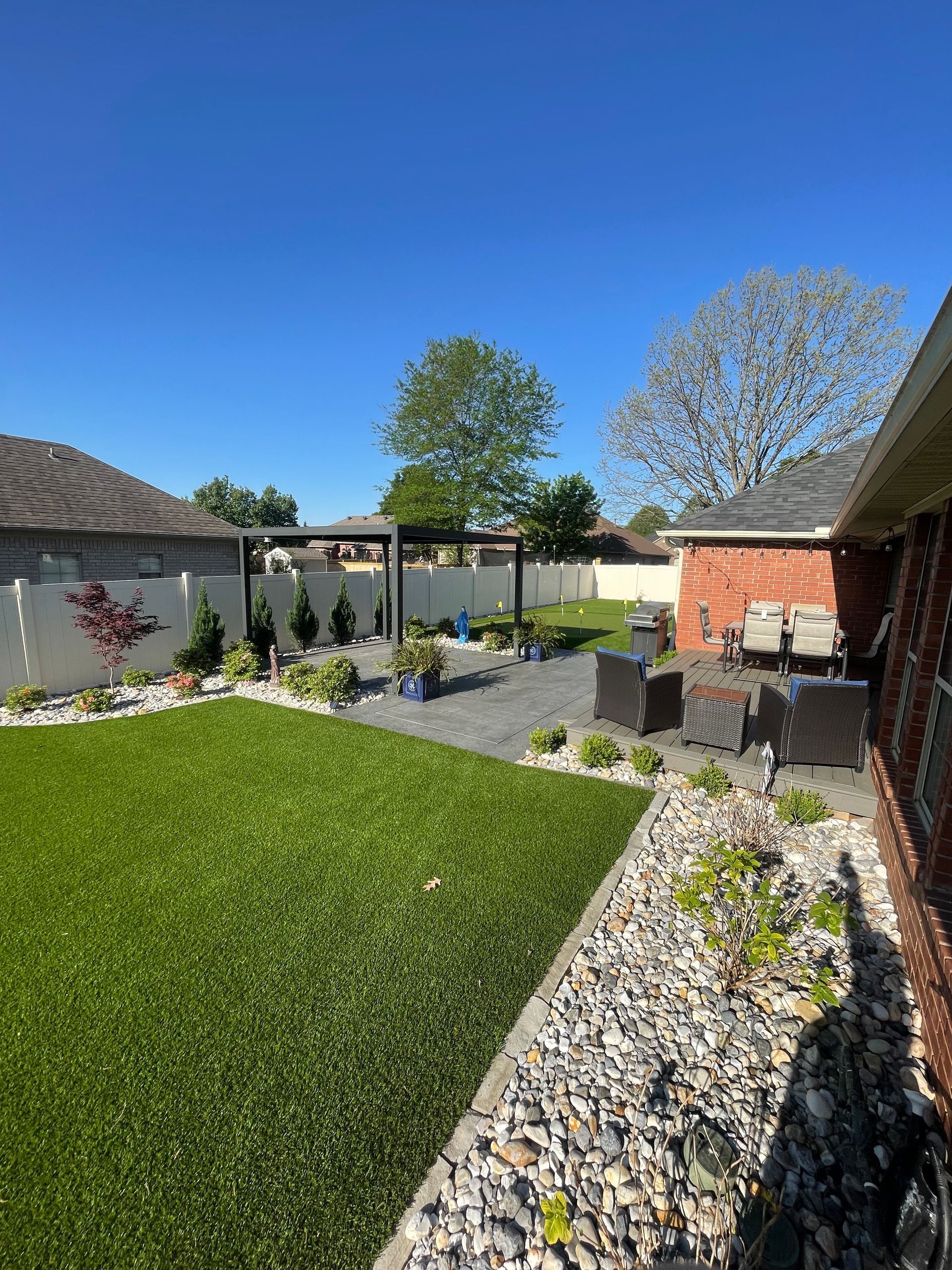Backyard with artificial grass, gravel patio, seating area, and landscaping under a blue sky.