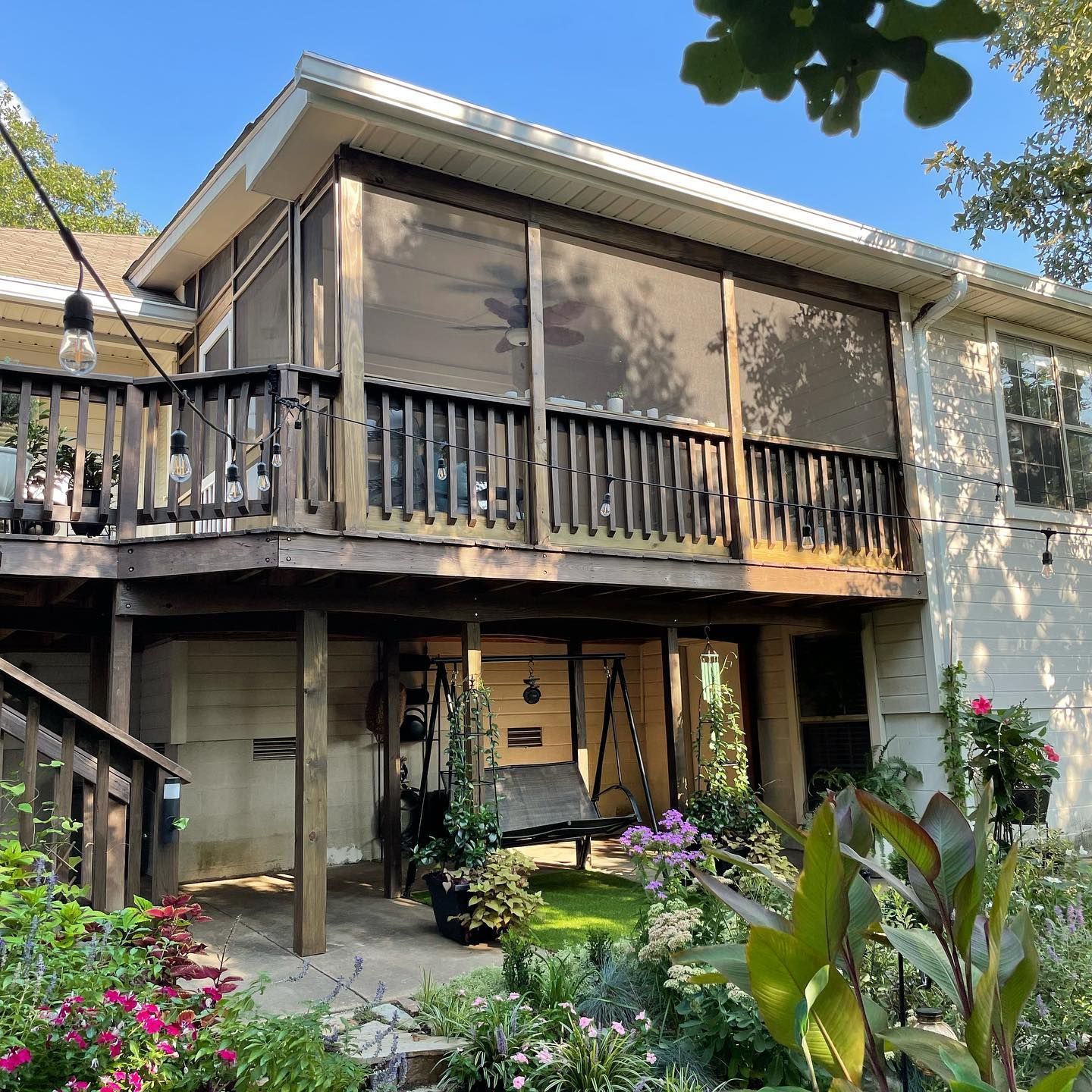 Wooden deck with screens and a lower level with a swing, surrounded by a garden with flowers.