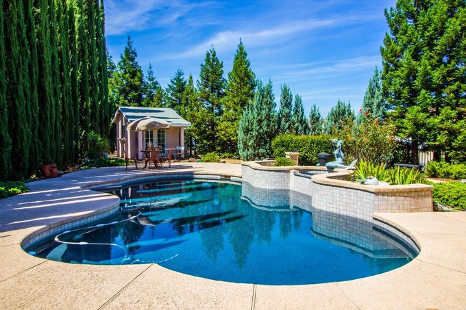 Backyard swimming pool with stone patio, trees, and a small gazebo under a blue sky