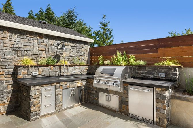 Outdoor stone patio with built-in grill and cabinets beside a wooden fence and stone wall