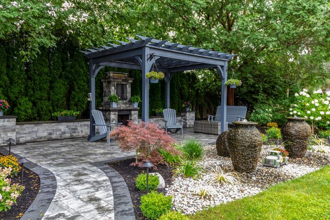 Stone patio garden with a dark pergola, potted plants, and landscaped shrubs under trees
