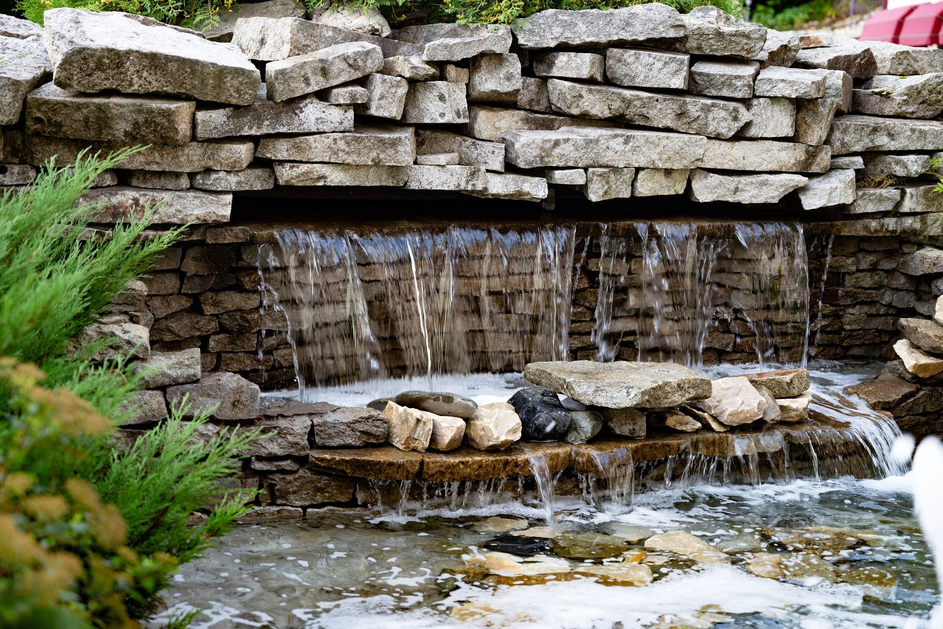 Stone waterfall cascading into a pond, with stacked rocks and greenery around it