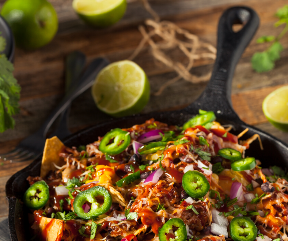 Pulled pork nachos in a cast iron skillet, topped with jalapeños, onions, and cilantro, with limes in the background.