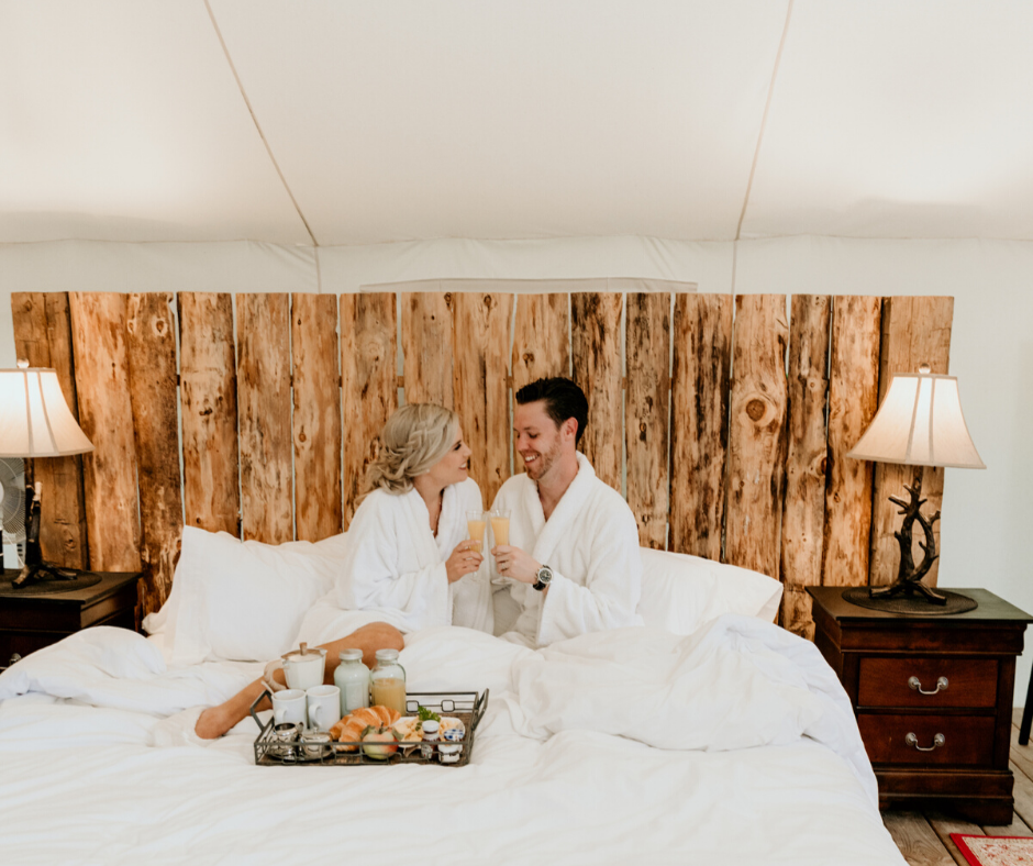Couple in white robes on a bed, toasting drinks. Tray of food between them. Rustic wood headboard.
