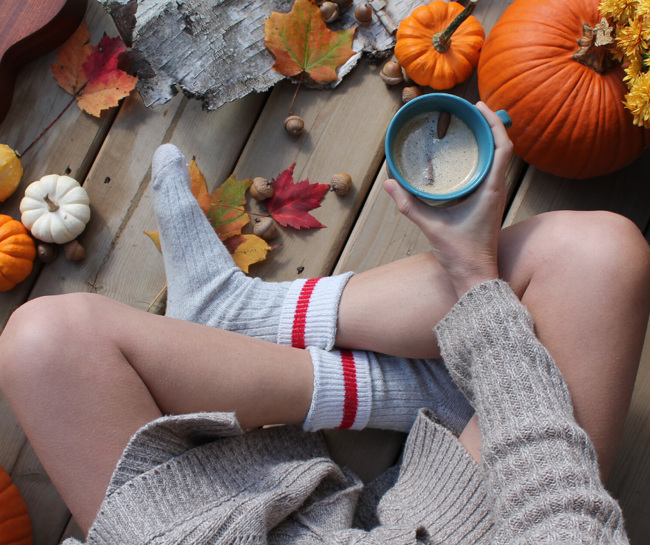 Person sitting outside in fall scene, holding coffee, wearing cozy sweater and socks with pumpkins and leaves.