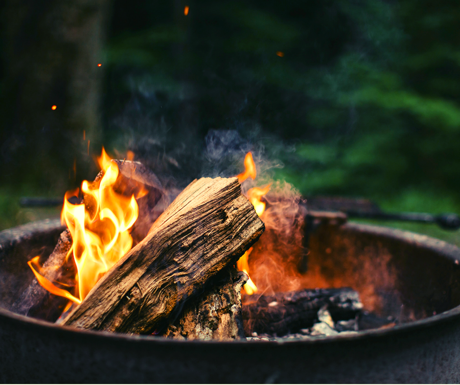 Campfire burning in a metal fire pit with flames and smoke, surrounded by trees.