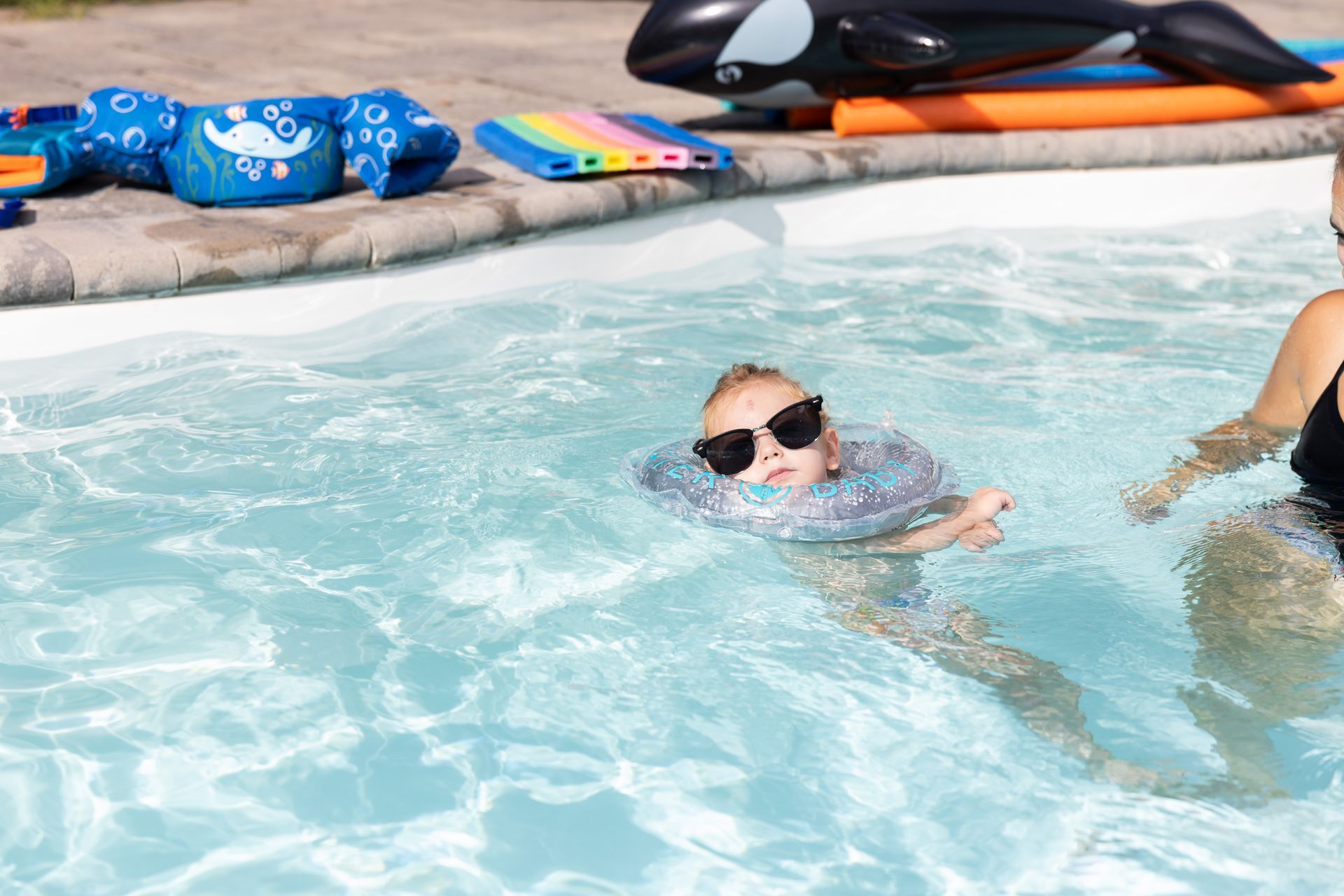 Child wearing sunglasses and a swim ring in a pool, supervised by an adult. Pool toys in background.