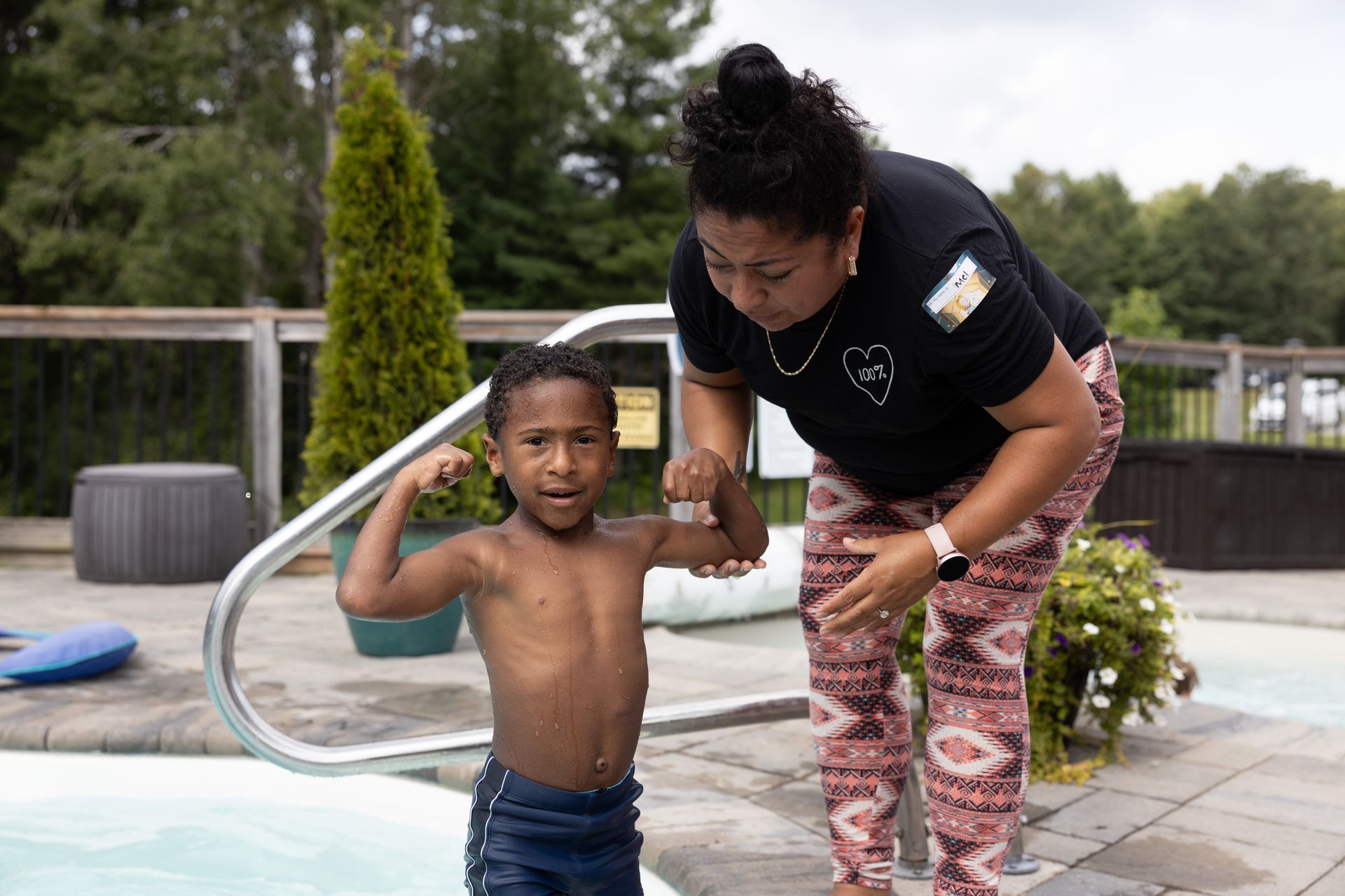 Child flexing bicep with adult, near pool. Adult wears black shirt and patterned pants, guiding child. Outdoors, sunny.
