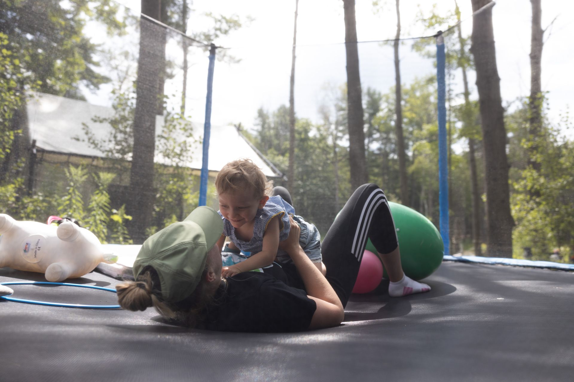 Woman on trampoline lifting a child, trees in background.