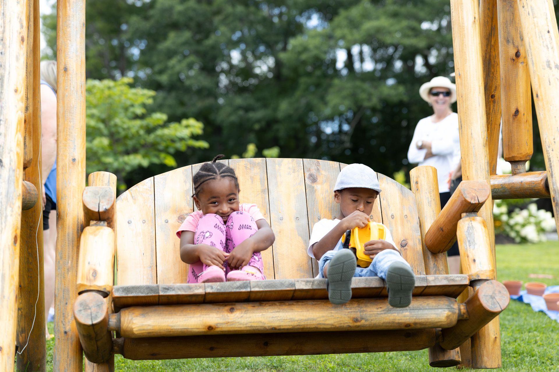 Two children sit on a wooden swing set. A woman watches in the background. Green grass and trees.