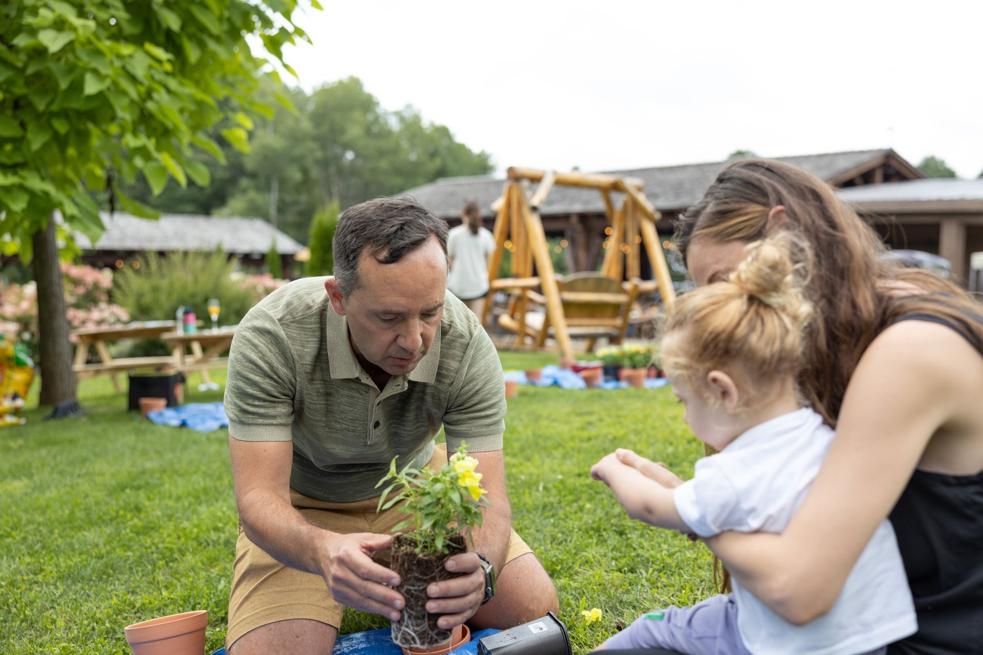 Family planting flowers together in a garden. Man holds plant, woman and child watch.