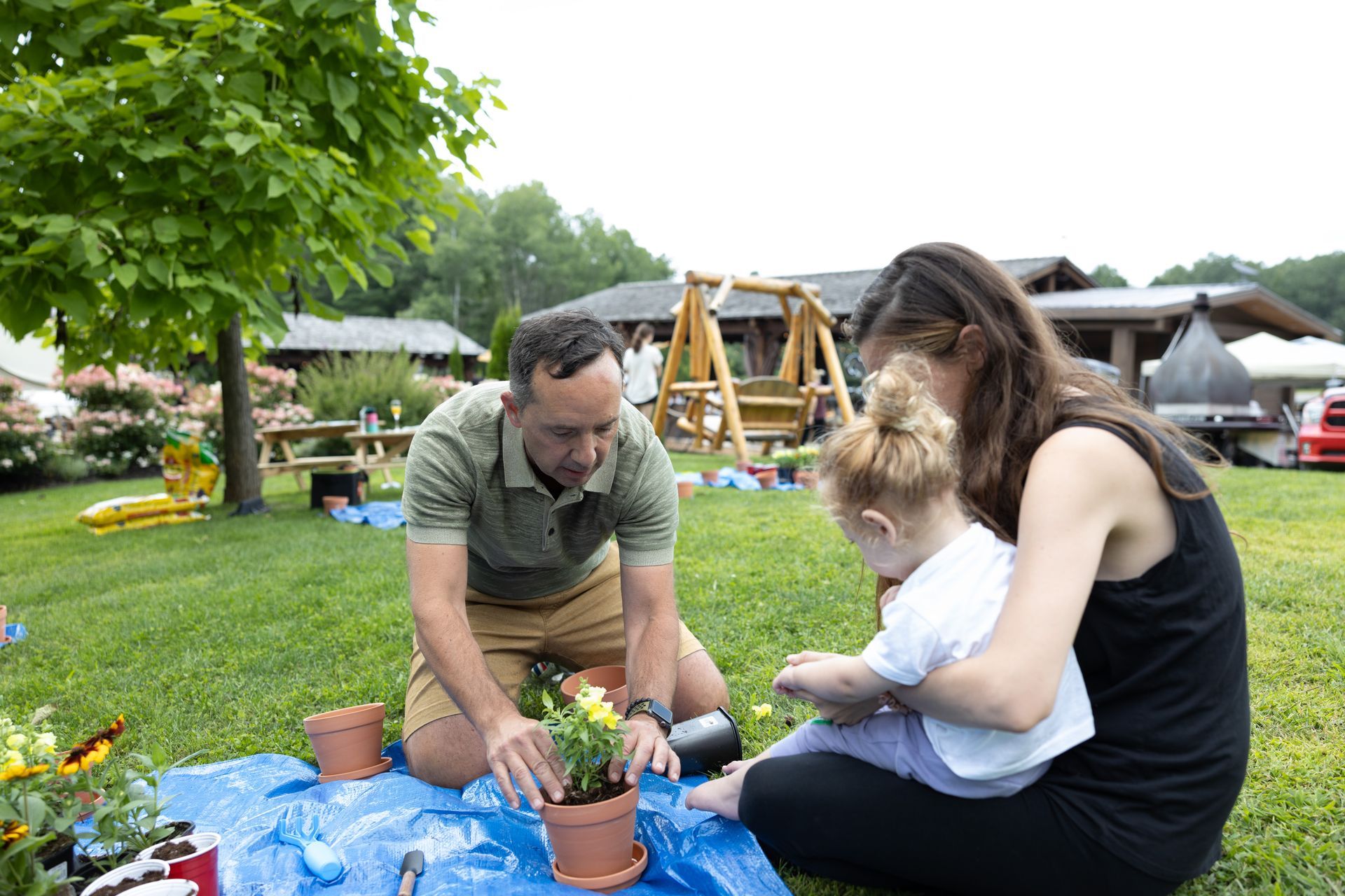 Family planting flowers outdoors; man kneeling, woman holding toddler.