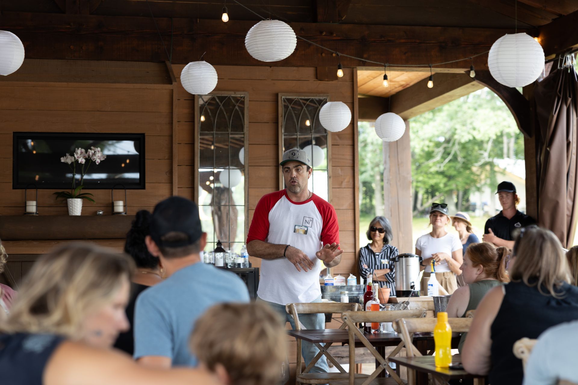 Man in red and white shirt speaks to a group under an outdoor canopy with string lights and paper lanterns.