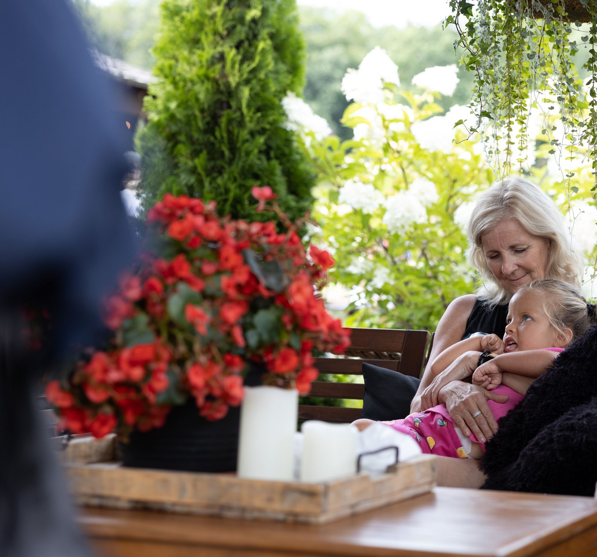 Woman and child on a patio, looking at something. Red flowers, candles on a tray, foliage in the background.