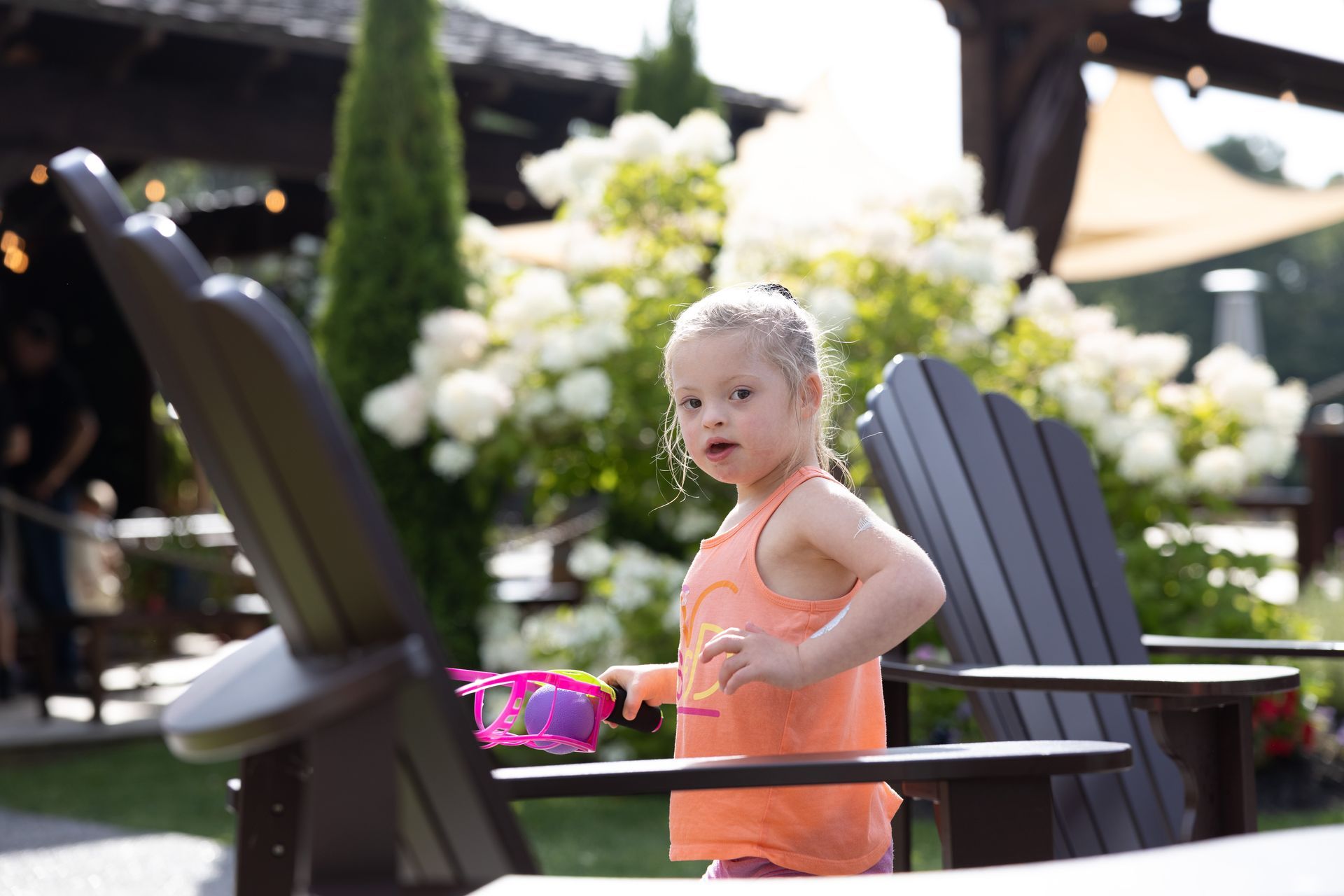 Girl with Down syndrome, orange top, playing with bubbles outside near chairs and flowers.